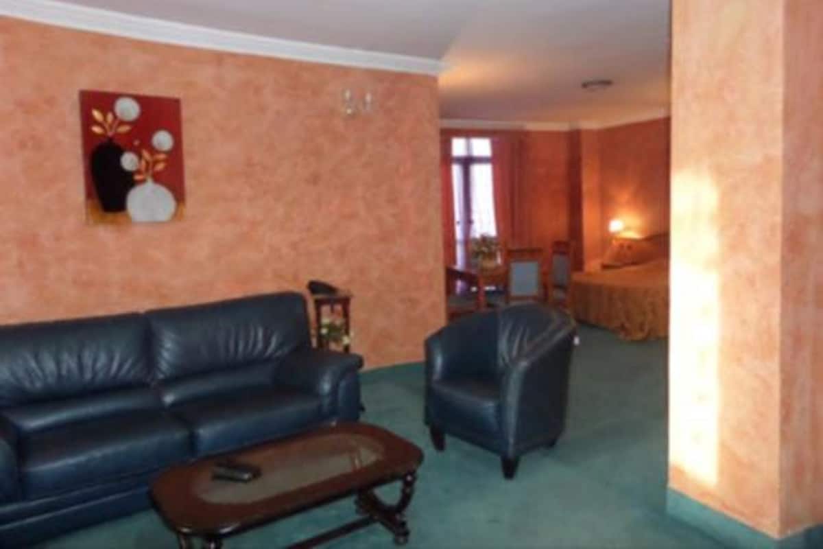 A living room with a dark blue sofa, a wooden coffee table, and a small armchair. There is a painting on the wall and a lamp on a side table.