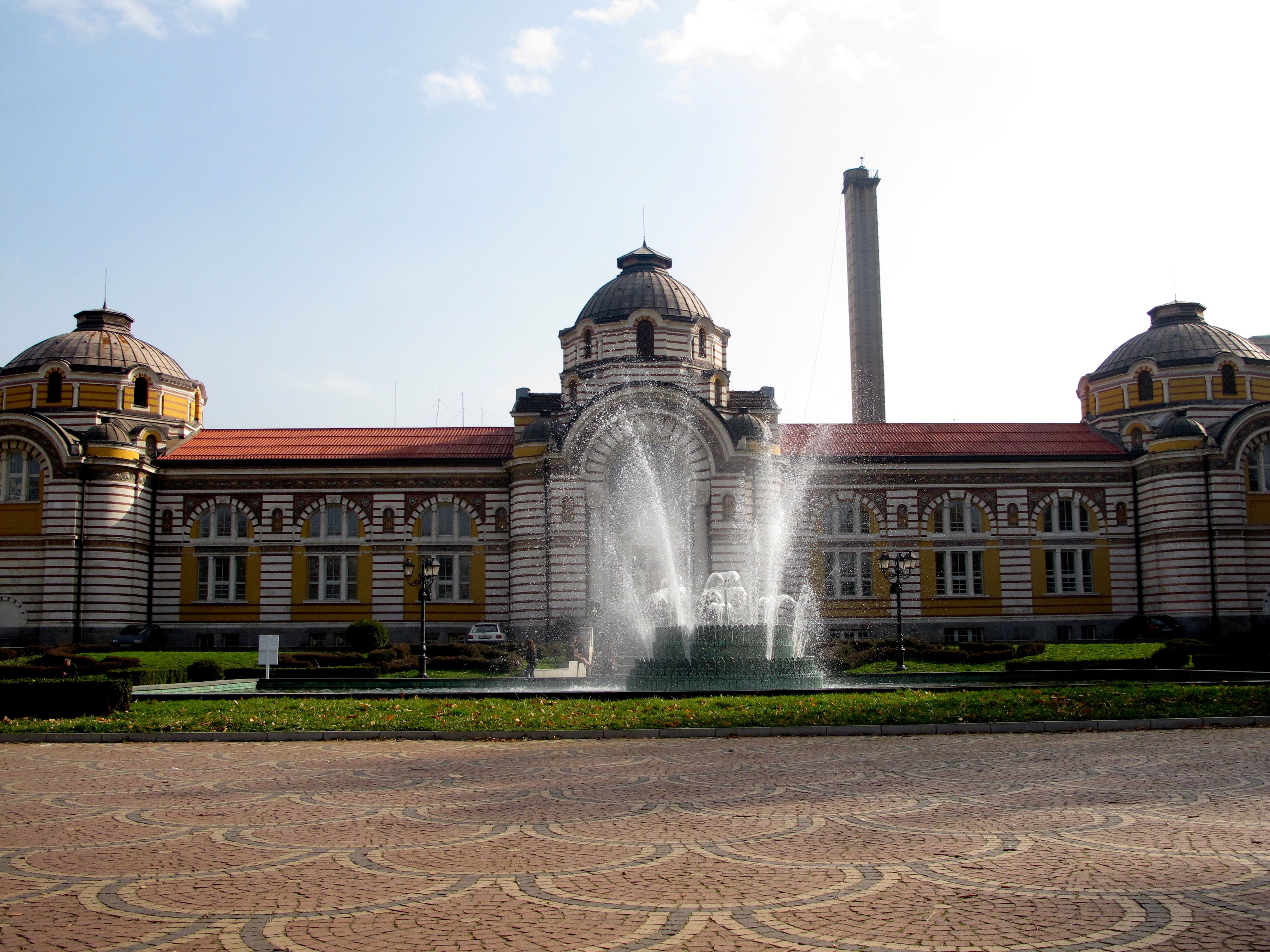 Un edificio histórico con una fuente central, cúpulas imponentes y una chimenea alta.