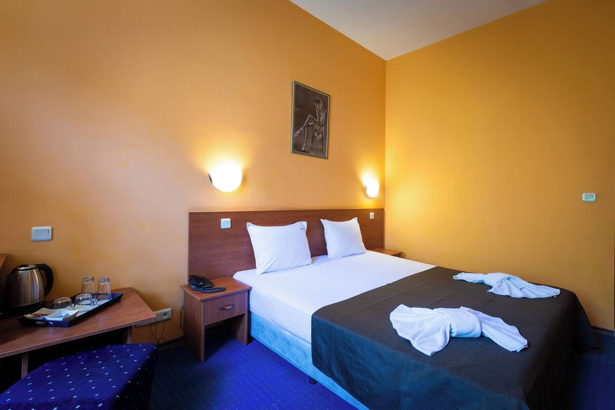 Habitación de hotel con cama, mesita de noche, lámpara y un pequeño escritorio con hervidor y vasos.