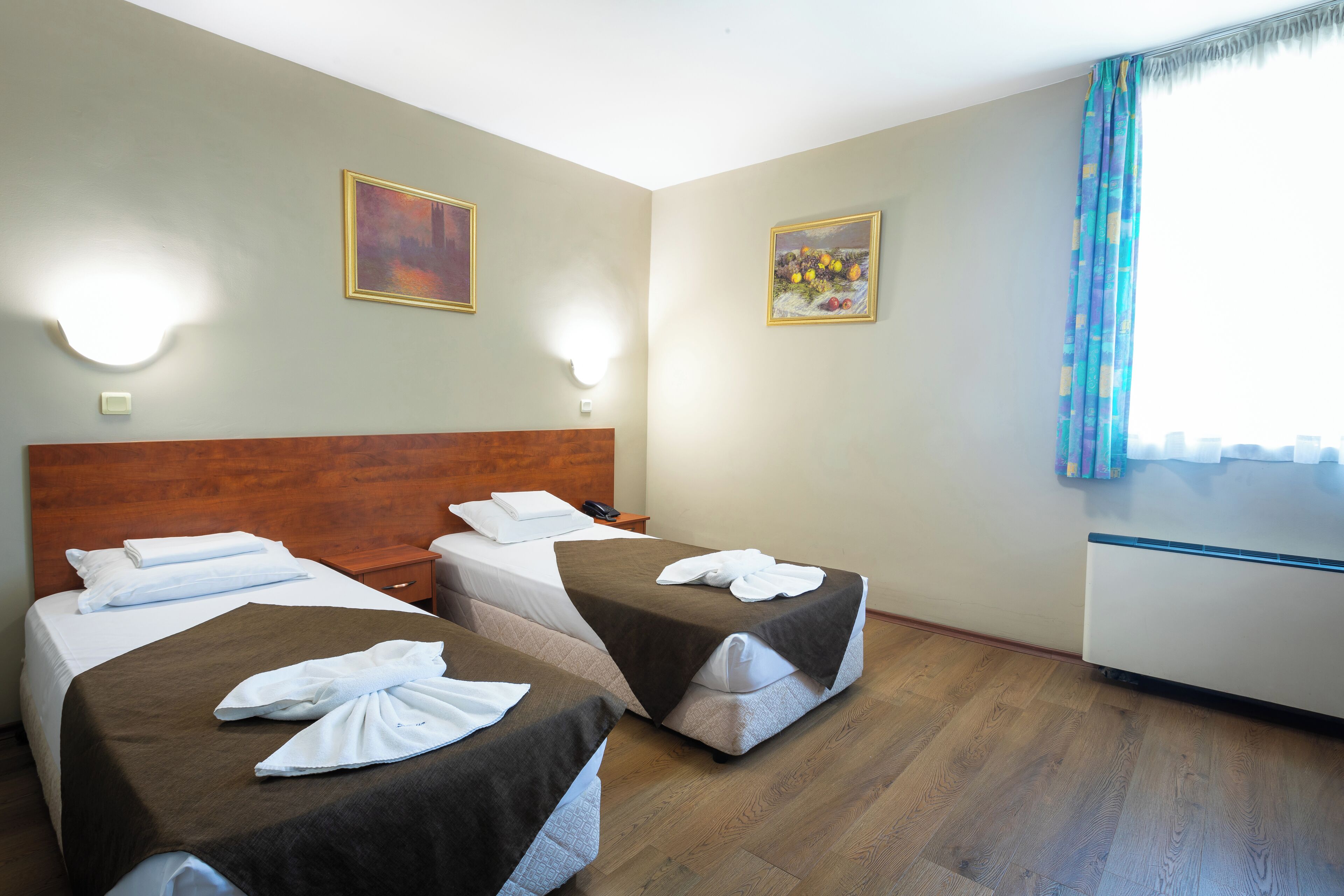 Une chambre d’hôtel avec deux lits, une tête de lit en bois, des lampes fixées au mur et une fenêtre avec des rideaux.