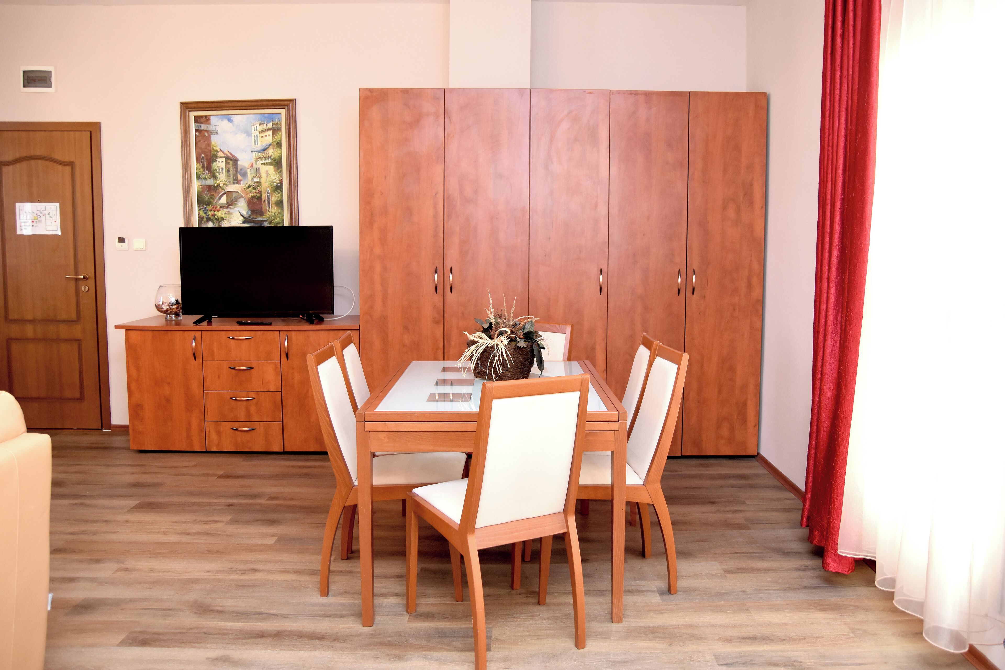Un comedor con una mesa de madera, sillas blancas, un televisor sobre un mueble de madera y un amplio armario.