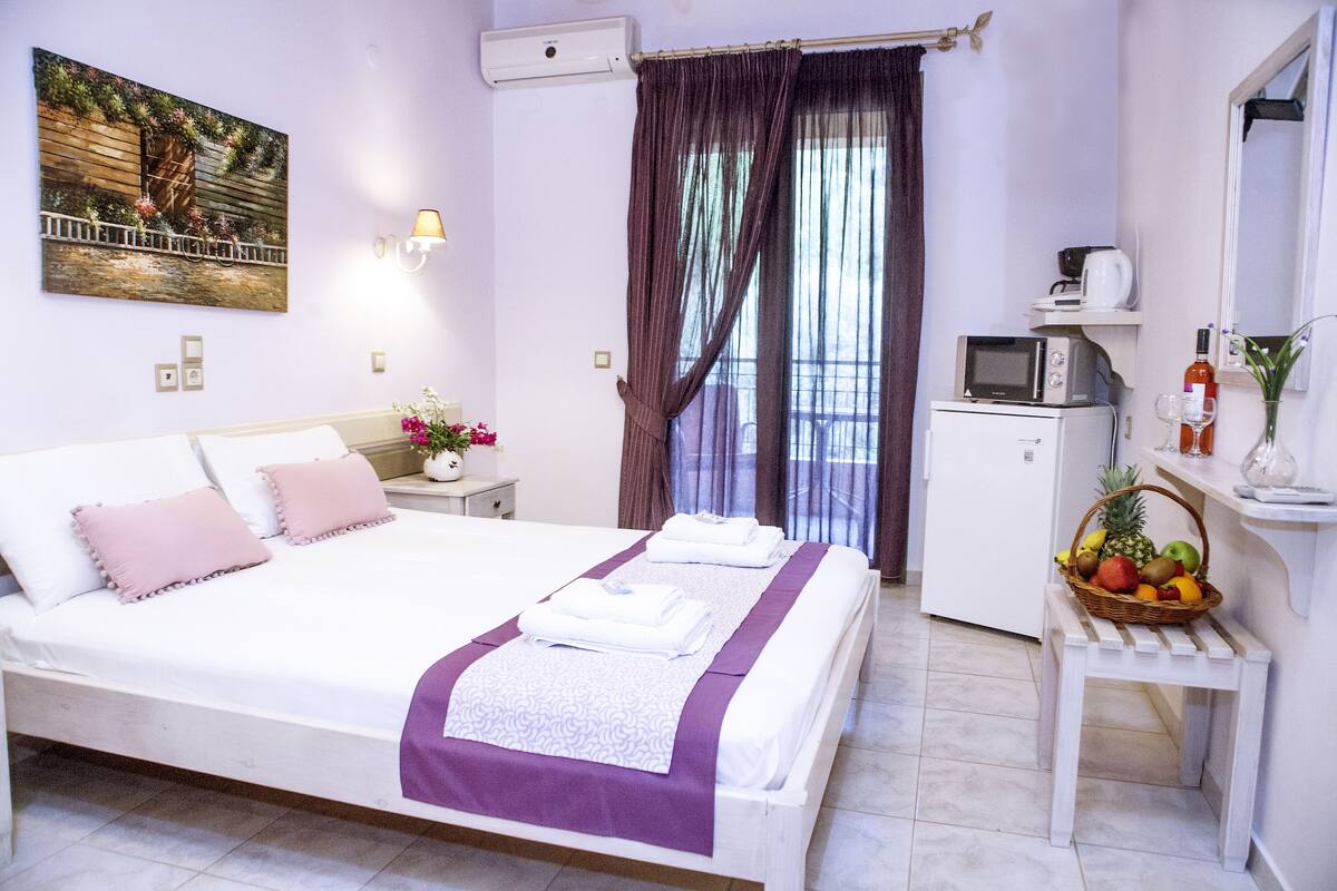A hotel room with a bed, a small kitchenette, a window with curtains, and a wall-mounted air conditioner.