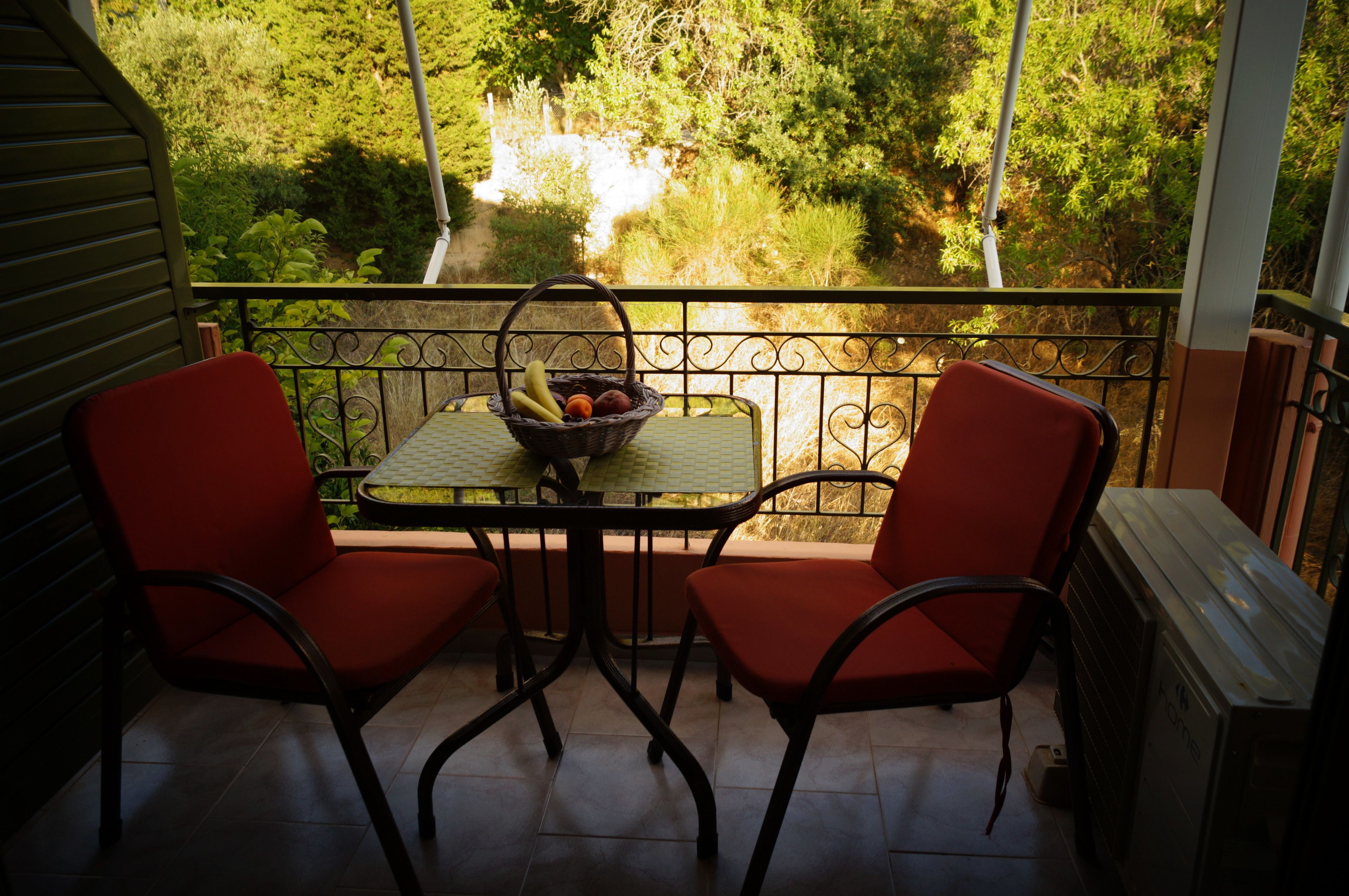 A balcony with two red chairs, a table with a fruit basket, and a view of greenery.