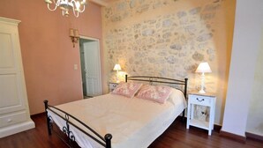 Un dormitorio con pared de piedra, una cama con colcha blanca, dos mesitas de noche con lámparas y una araña.