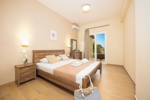 A hotel room with a wooden bed, a nightstand, a dresser, a mirror, and a sliding glass door to the balcony. - Koulouris Beach (Corfu)