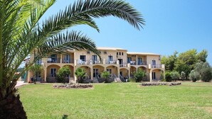 A multi-story building with a symmetrical facade, multiple balconies, and a well-maintained lawn. - Koulouris Beach (Corfu)