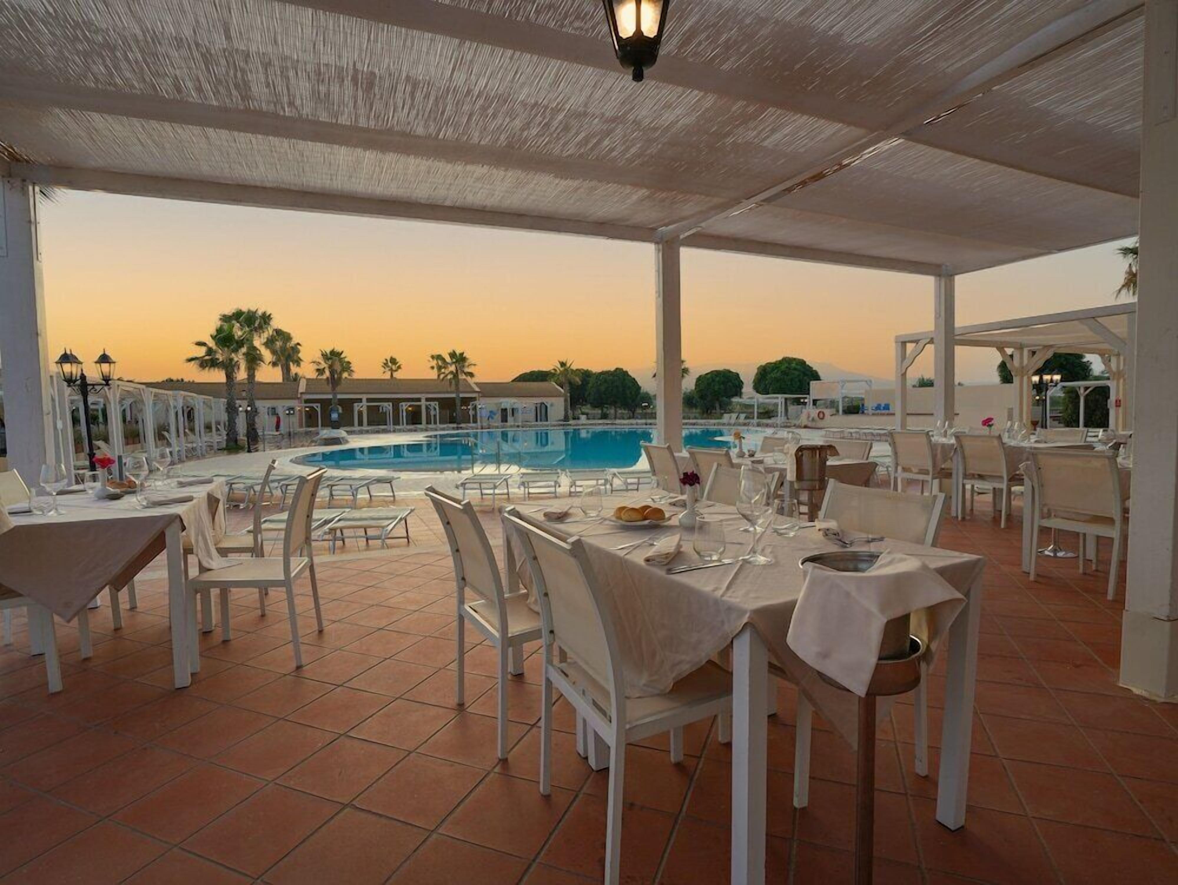 Zona pranzo all'aperto con tavoli e sedie, una piscina e vista sul tramonto.