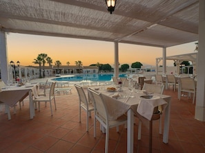 Un área de comedor al aire libre con mesas y sillas, una piscina y vistas al atardecer.