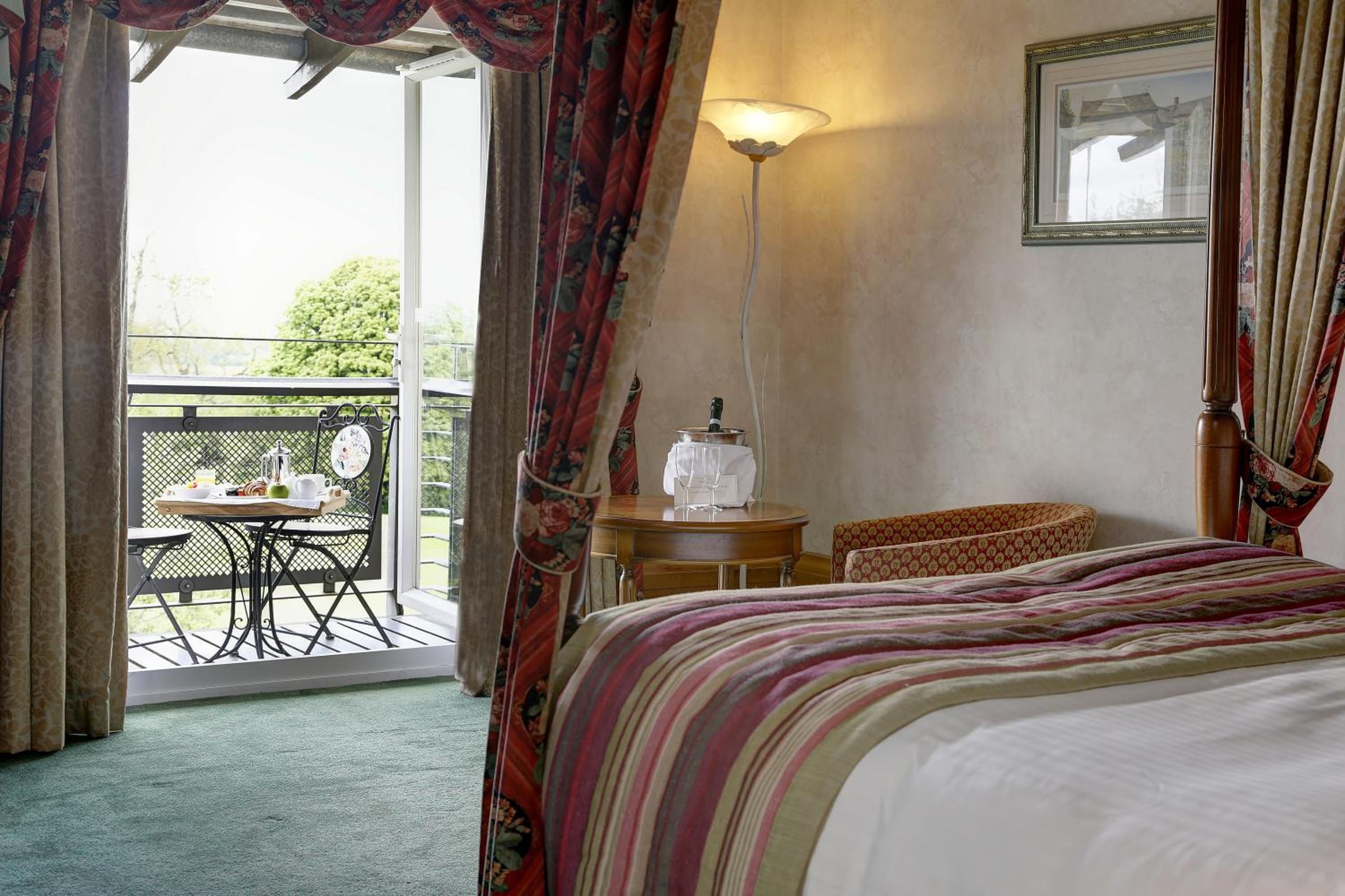 A hotel room with a bed, a table with a chair, a lamp, and a balcony with a table and chairs.