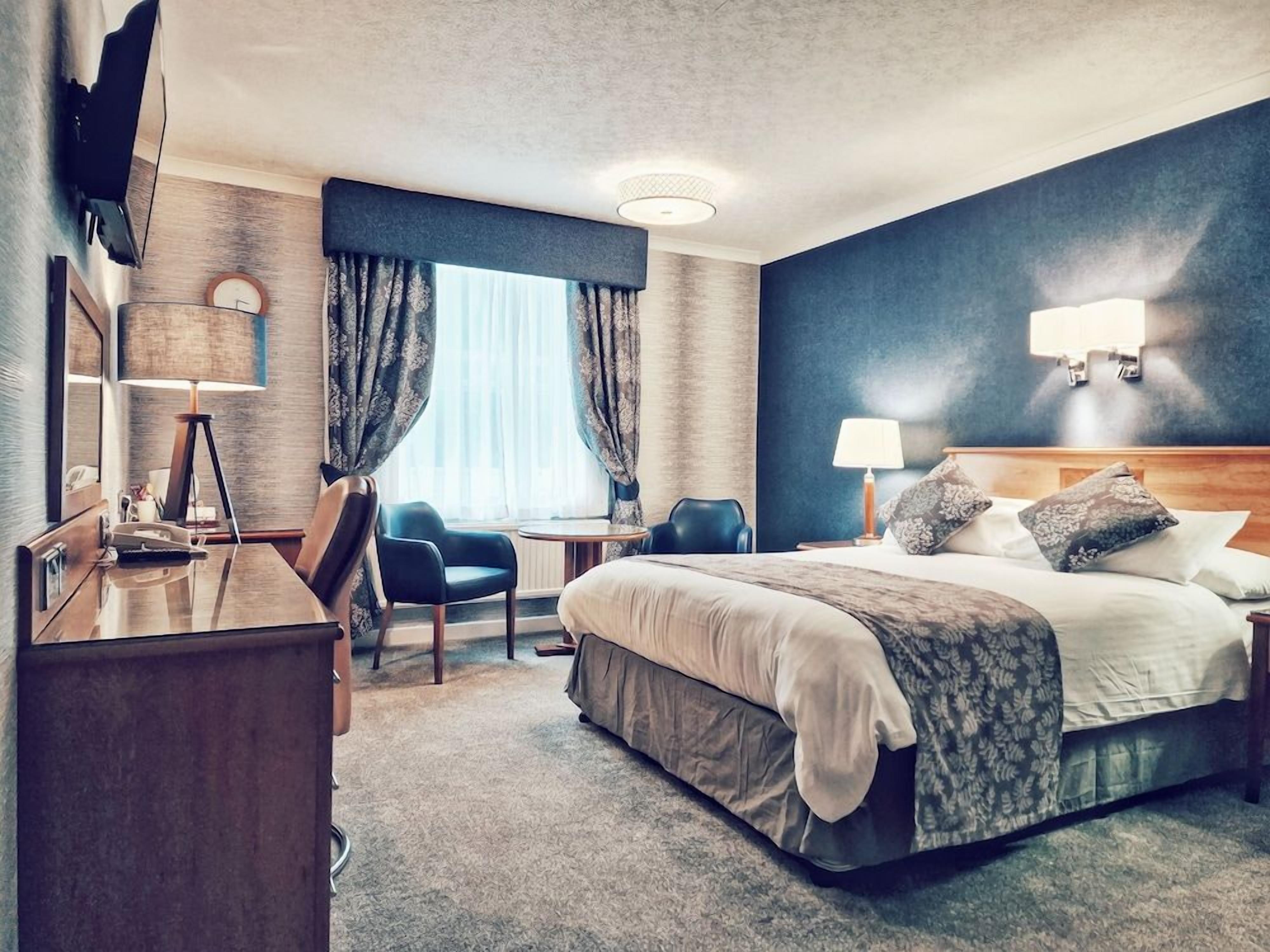 A hotel room with a bed, desk, chair, and a large window with curtains.
