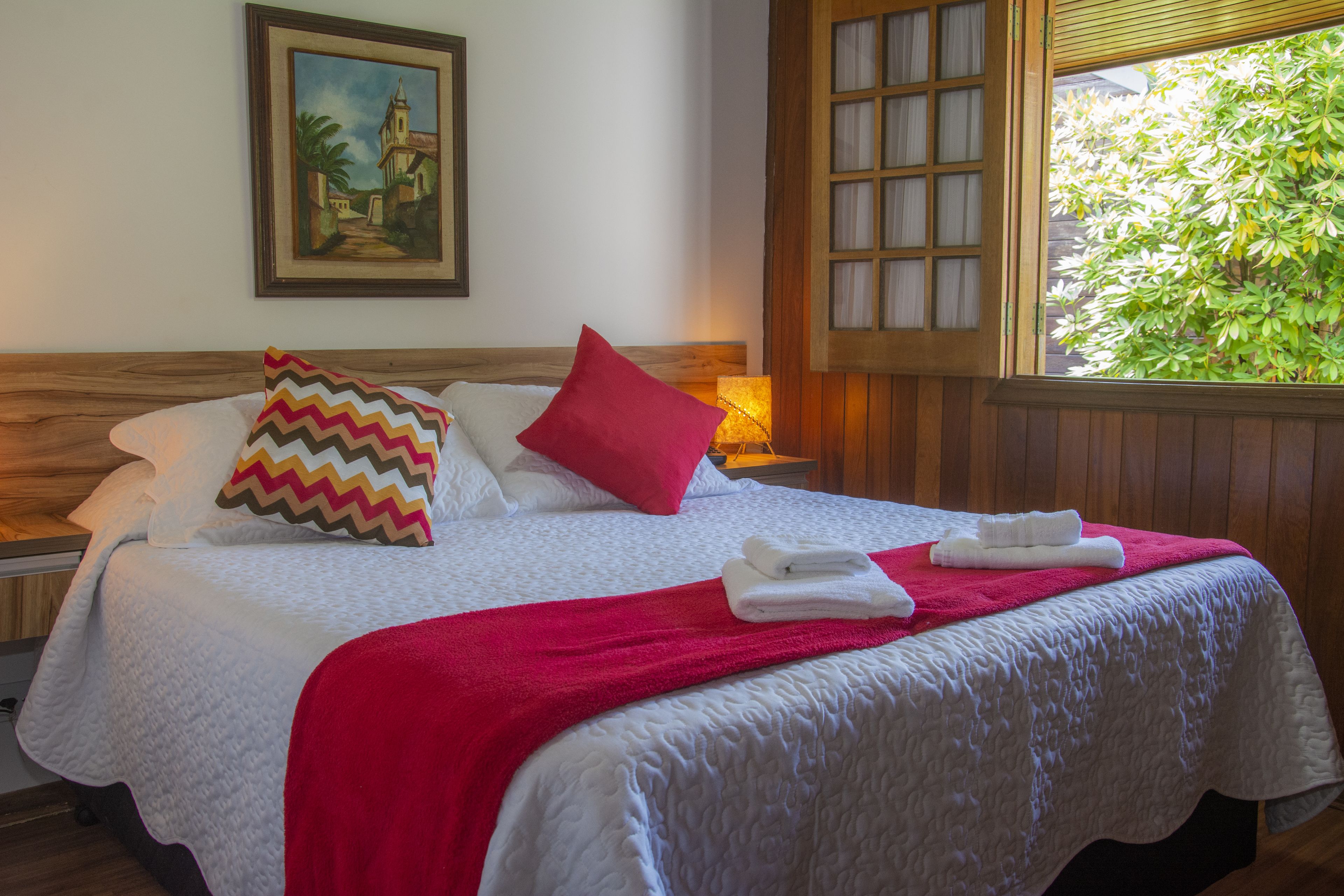 A neatly made bed with pillows, a red blanket, and towels, a painting on the wall, and a window with a view of greenery.