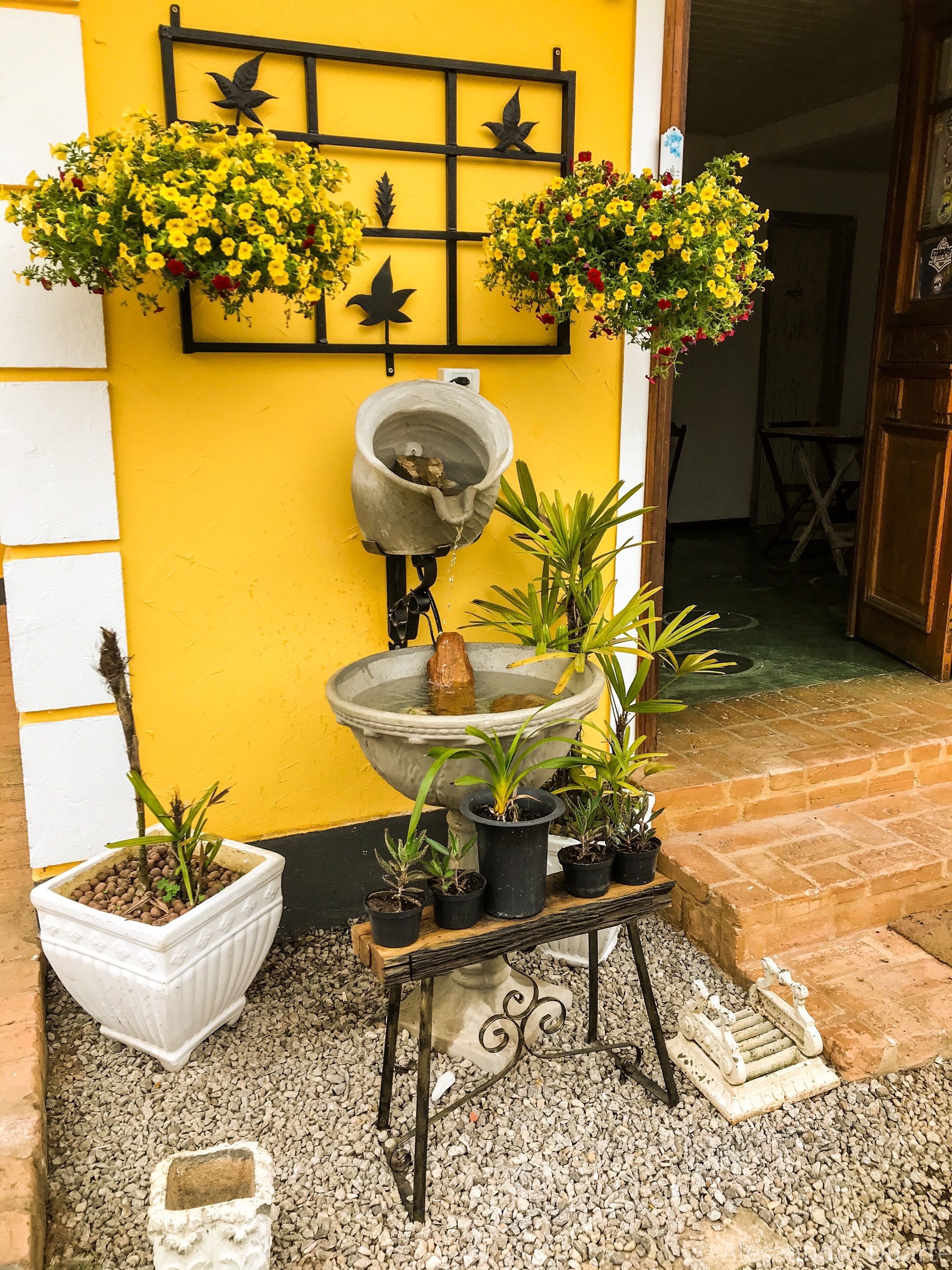 A courtyard with a yellow wall, a bird-shaped metal decoration, a large planter with yellow flowers, and a small fountain.