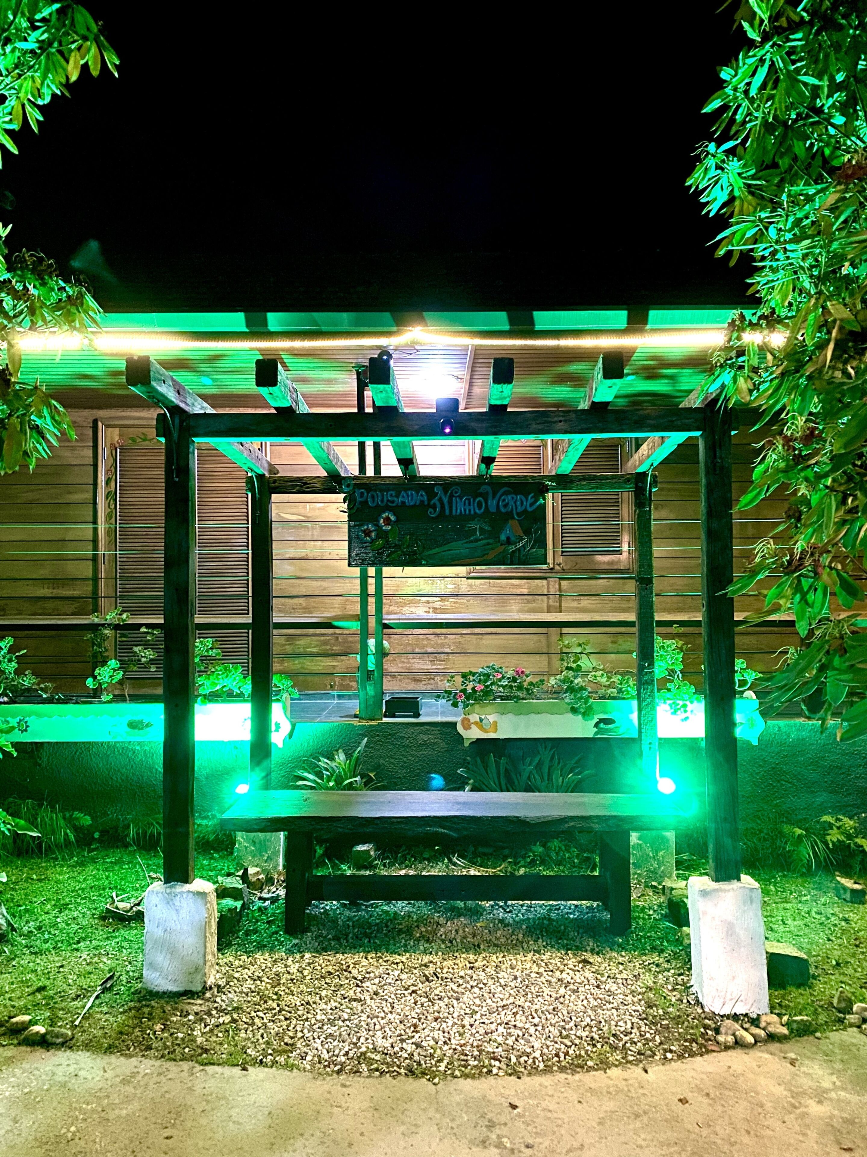 Un belvédère surmonté d’une enseigne indiquant « Pousada Nino Verde », la nuit, entouré de verdure et illuminé par des lumières vertes.