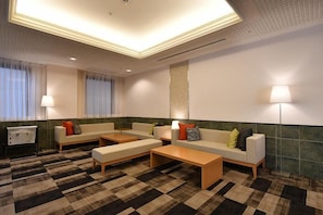 Vestíbulo de hotel con mostrador de recepción, zona de estar con sofás y una mesa de centro, y un ventanal con cortinas.