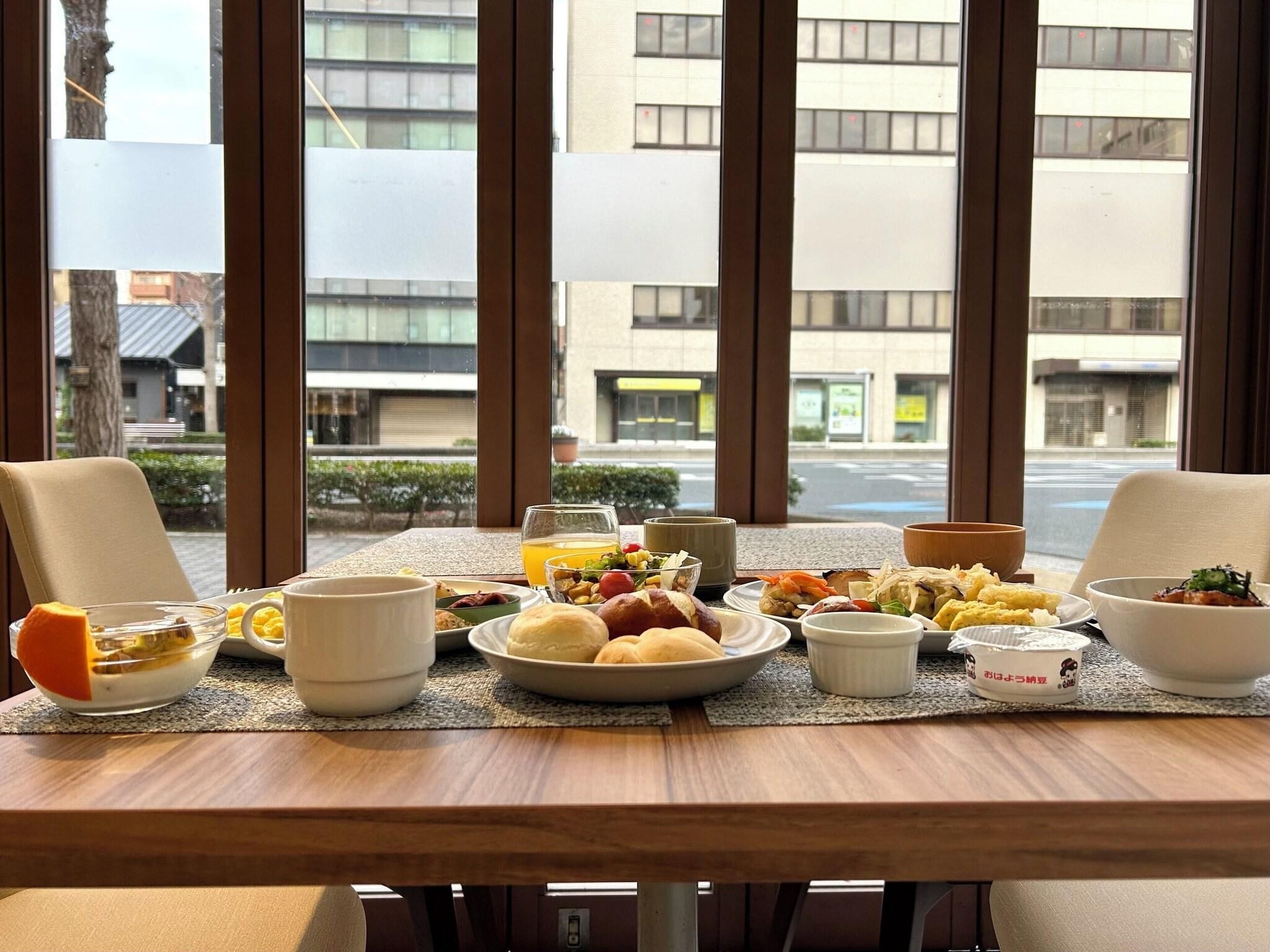 Une table de petit-déjeuner avec différents plats, un panier de fruits et une tasse, le tout placé devant une fenêtre donnant sur des immeubles.