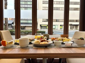 Une table de petit-déjeuner avec différents plats, un panier de fruits et une tasse, le tout placé devant une fenêtre donnant sur des immeubles.