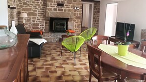 Un salon avec un mur en pierre, une cheminée, des fauteuils verts, une table à manger en bois et un téléviseur.
