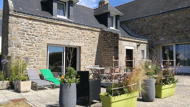 Une maison en pierre avec un toit en pente, de grandes fenêtres et un patio aménagé avec des meubles d’extérieur et des jardinières.