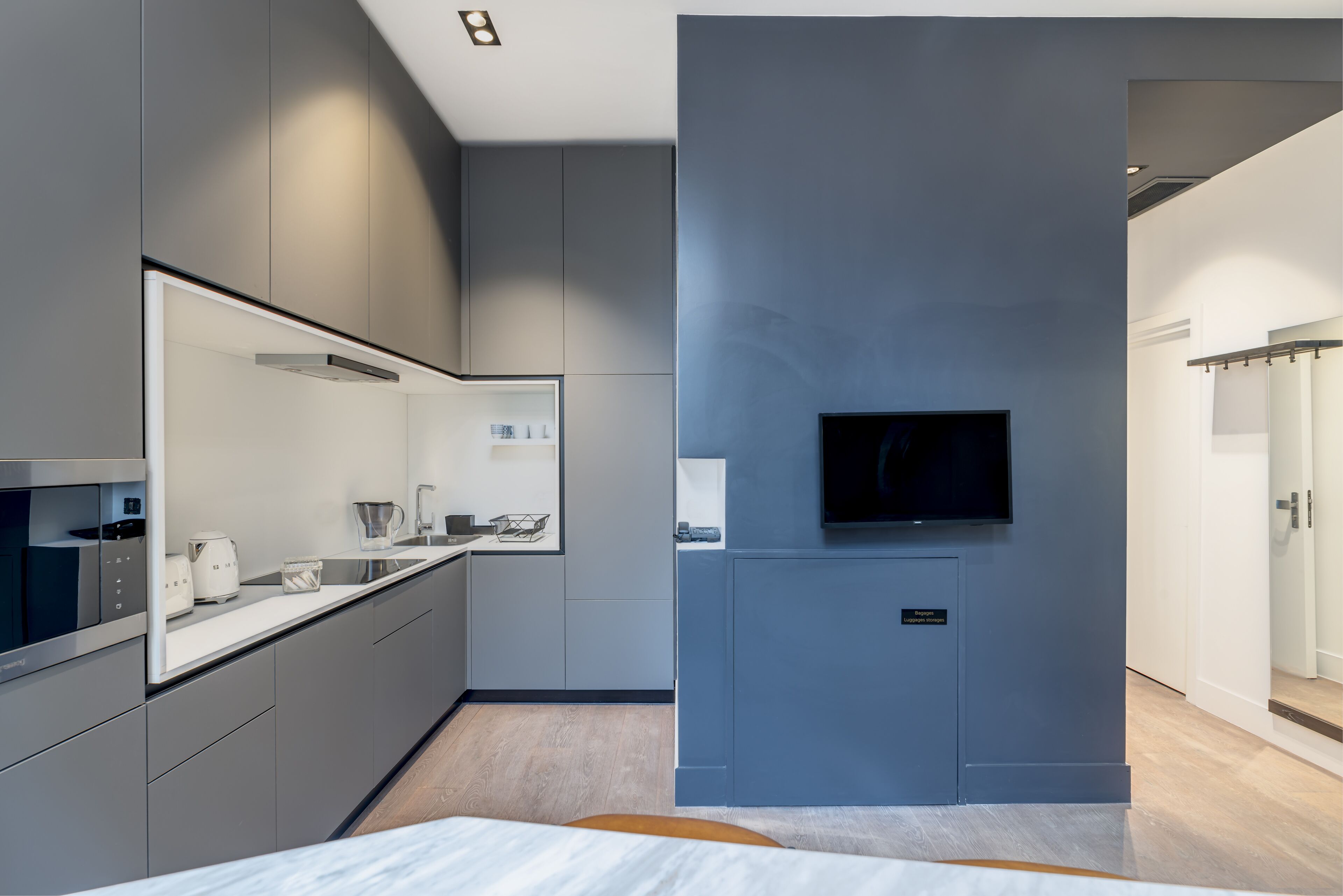 A modern kitchen with grey cabinetry, a built-in oven, and a flat-screen TV mounted on the wall.
