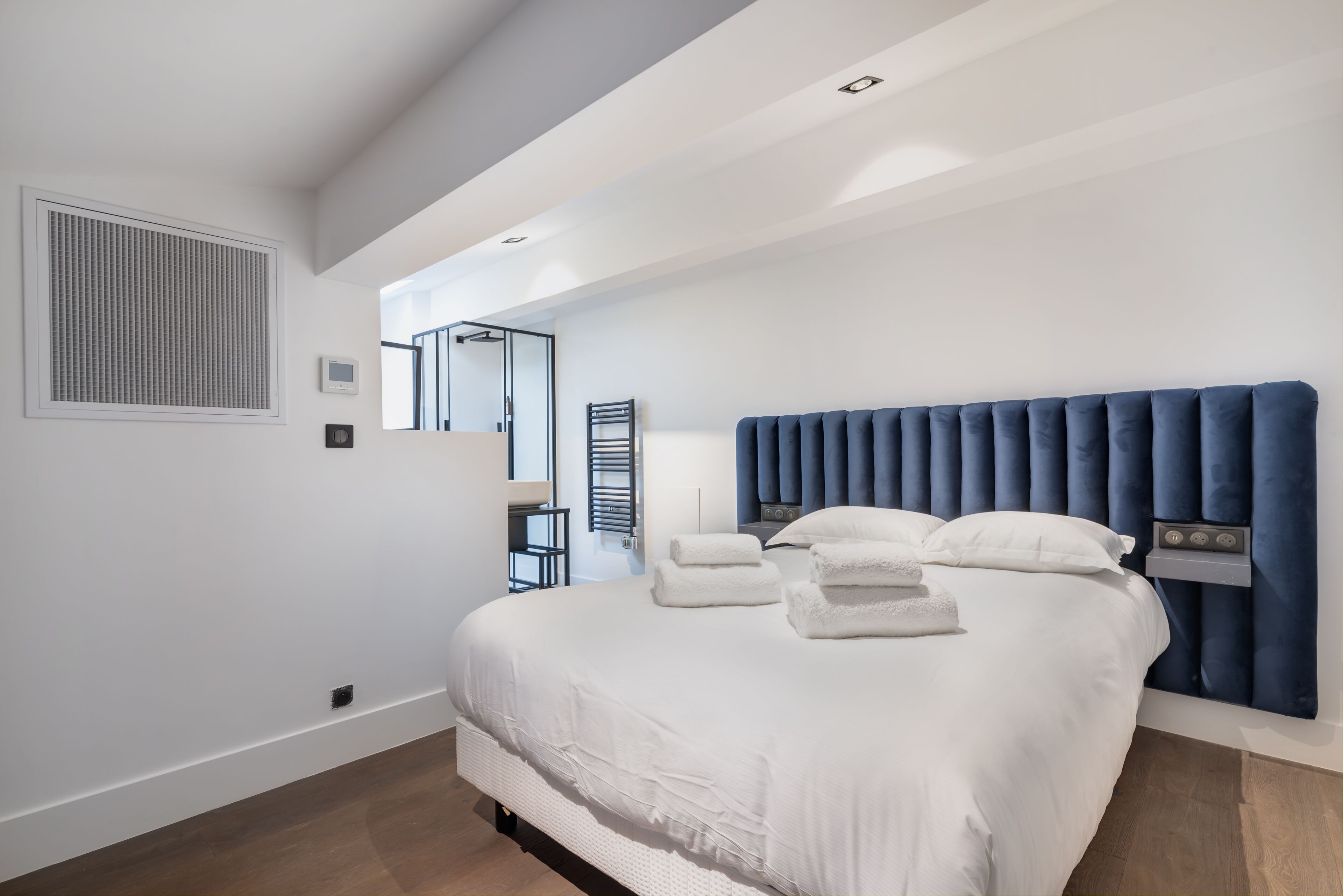 A modern bedroom with a large bed, white bedding, a blue headboard, and a view of a balcony.