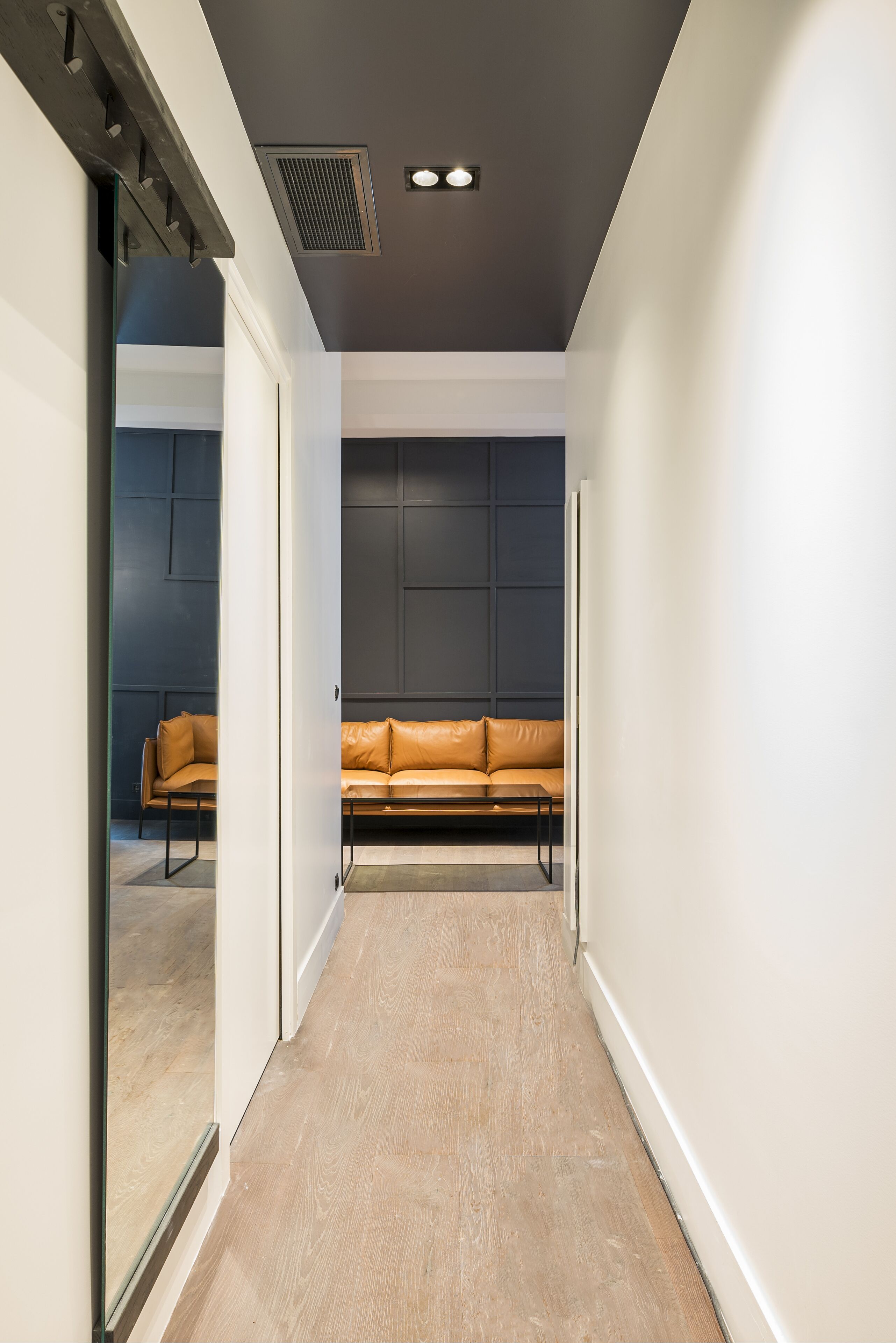 Um corredor com sofá de couro, piso de madeira e parede escura com design em painel.