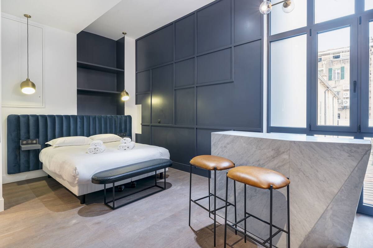 A modern hotel room with a bed, bench, two bar stools, a marble counter, and a large window offering a city view.