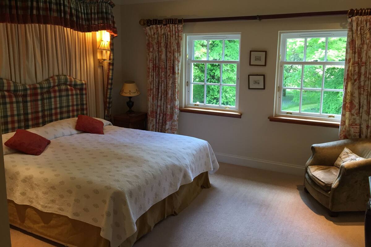 A bedroom with a bed, two windows, a chair, and a nightstand with a lamp.