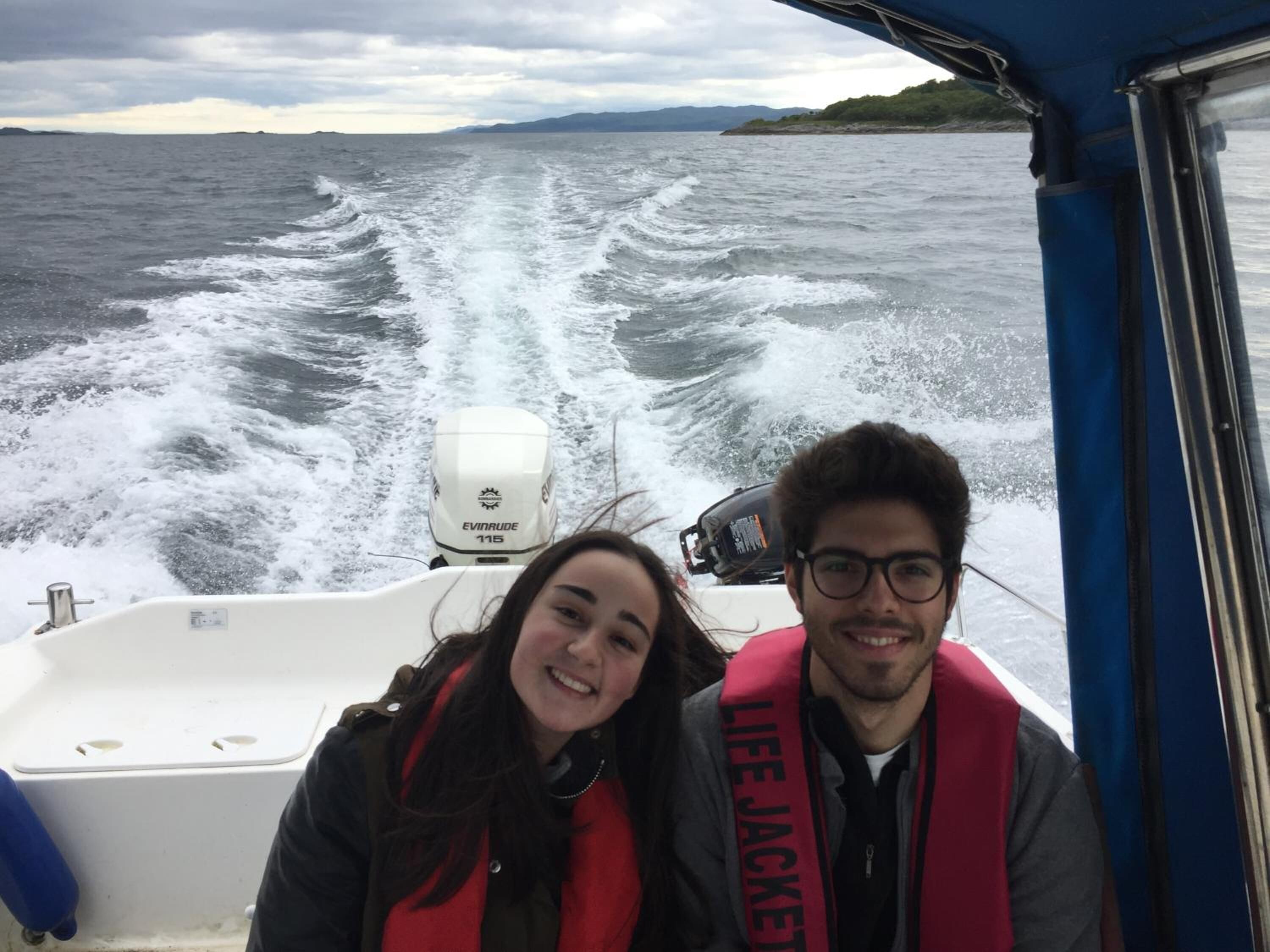 Un bote con dos personas usando chalecos salvavidas, un motor blanco y vista al mar y la costa lejana.