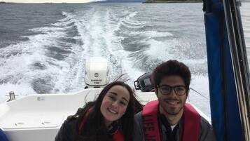 Un bote con dos personas usando chalecos salvavidas, un motor blanco y vista al mar y la costa lejana.