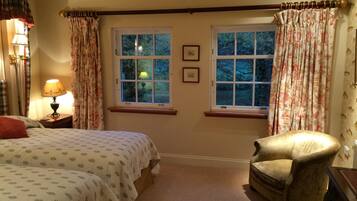 A cozy bedroom with two windows, a bed with patterned bedding, a chair, and a lamp.