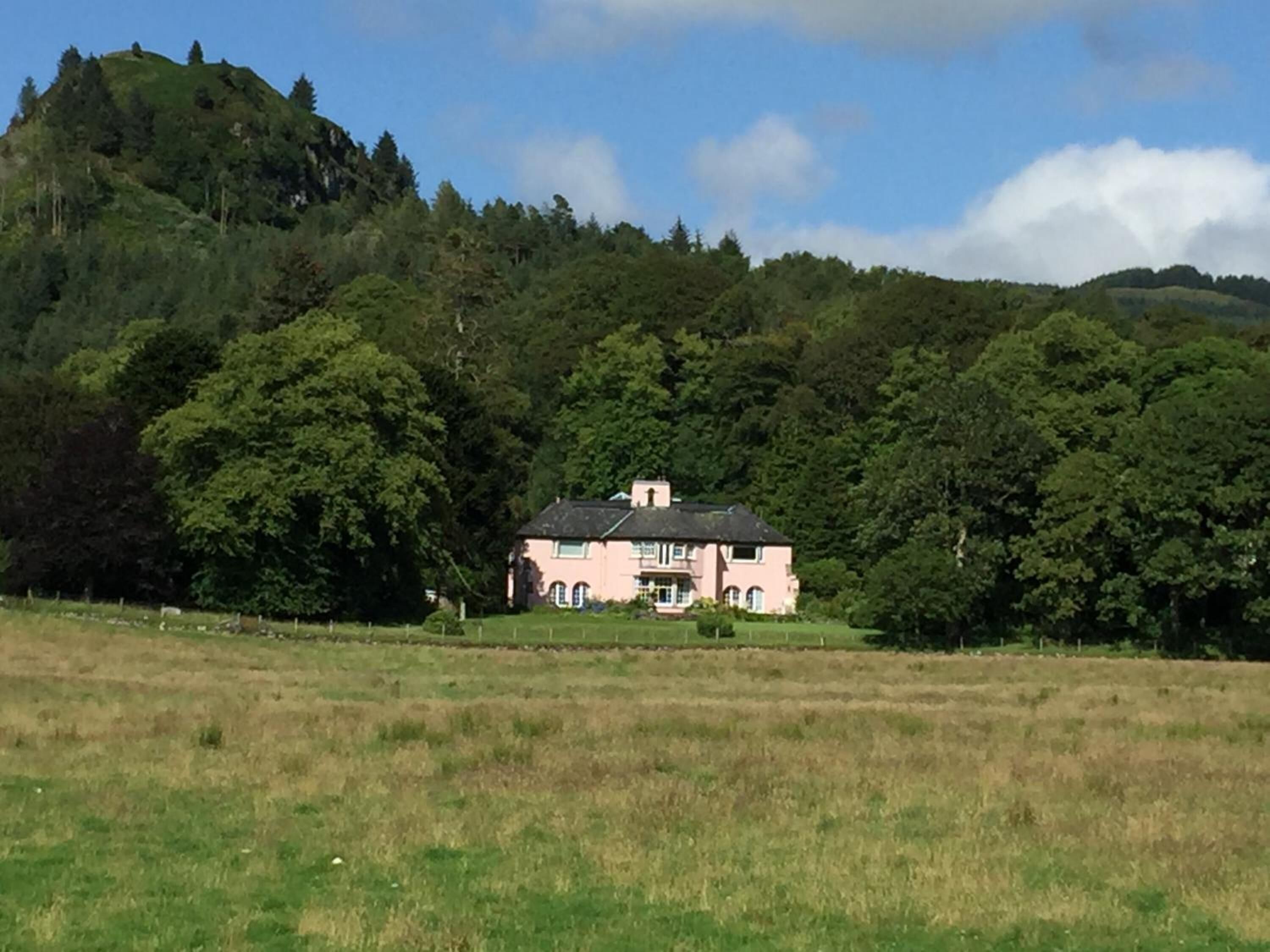 A pink house with a symmetrical facade, surrounded by a lush green landscape and a hill in the background.
