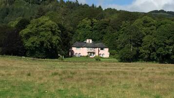 A pink house with a symmetrical facade, surrounded by a lush green landscape and a hill in the background.