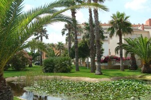 Un jardín con palmeras, un estanque cubierto de lirios y un edificio al fondo.