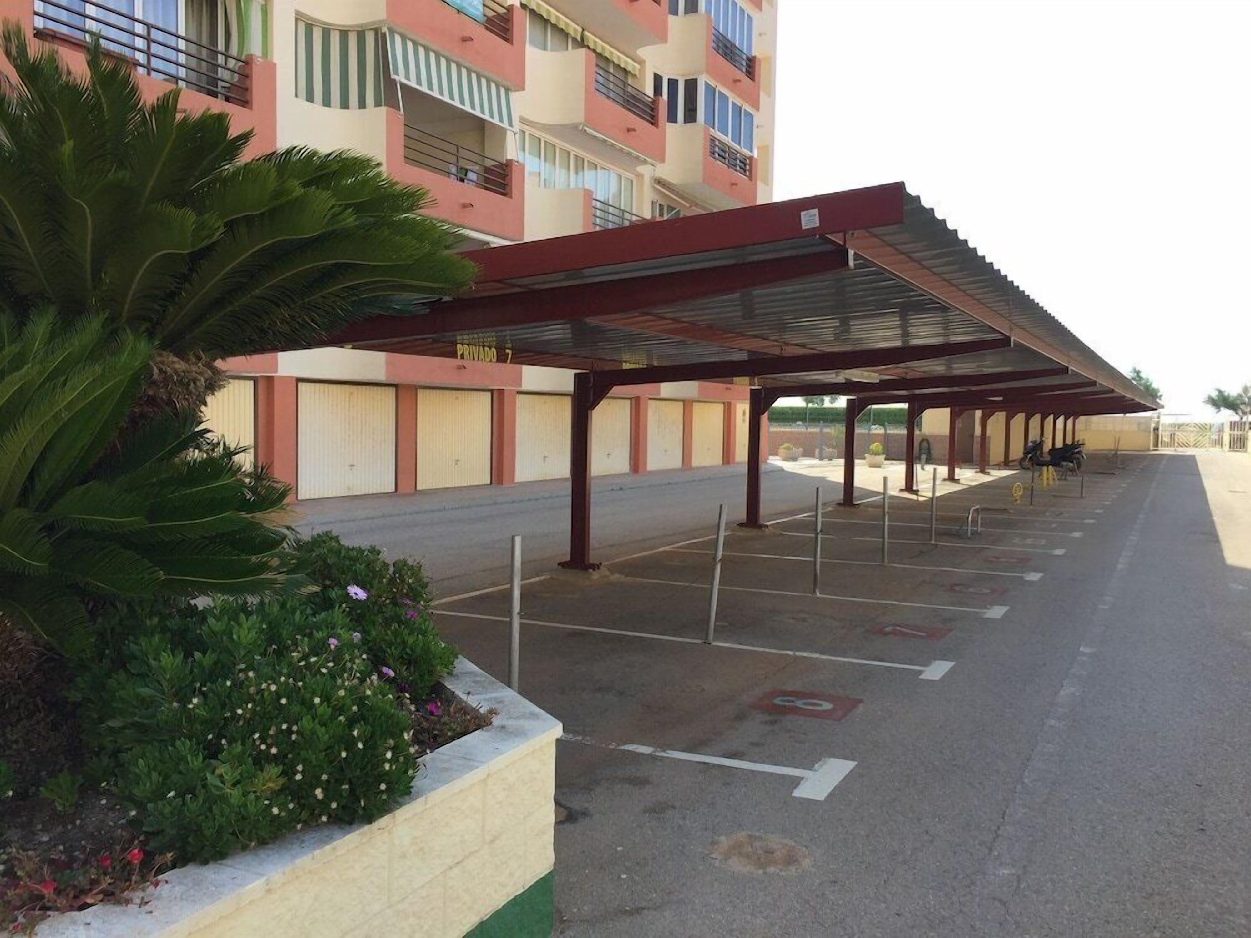 A covered parking area with a row of empty parking spaces, a building with balconies, and a row of plants.