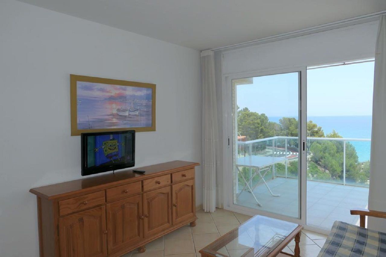 Una habitación con televisor sobre un mueble de madera, balcón con barandilla de vidrio y vista al mar.