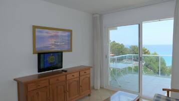 Una habitación con televisor sobre un mueble de madera, balcón con barandilla de vidrio y vista al mar.