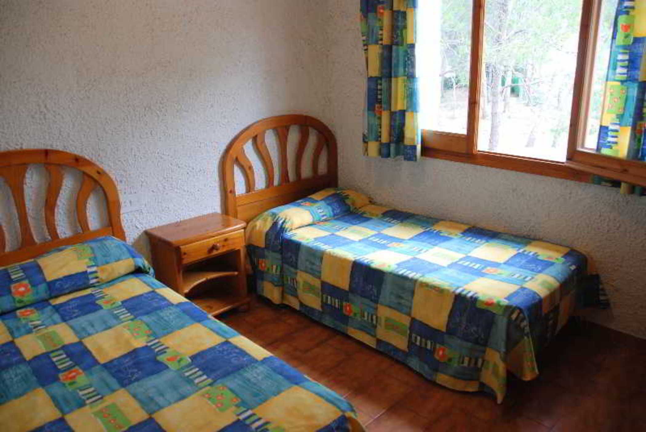 Une chambre avec deux lits en bois, chacun recouvert d’une courtepointe matelassée. Une table de chevet en bois se trouve entre les deux lits, et une fenêtre avec des rideaux est située sur le mur de droite.