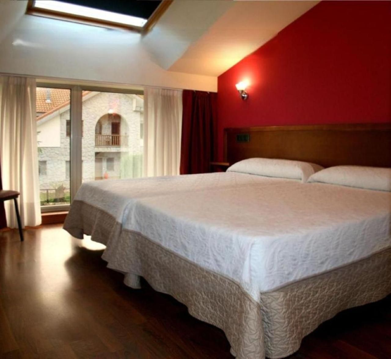 A hotel room with a large bed, a wooden floor, a red wall, and a window with curtains.