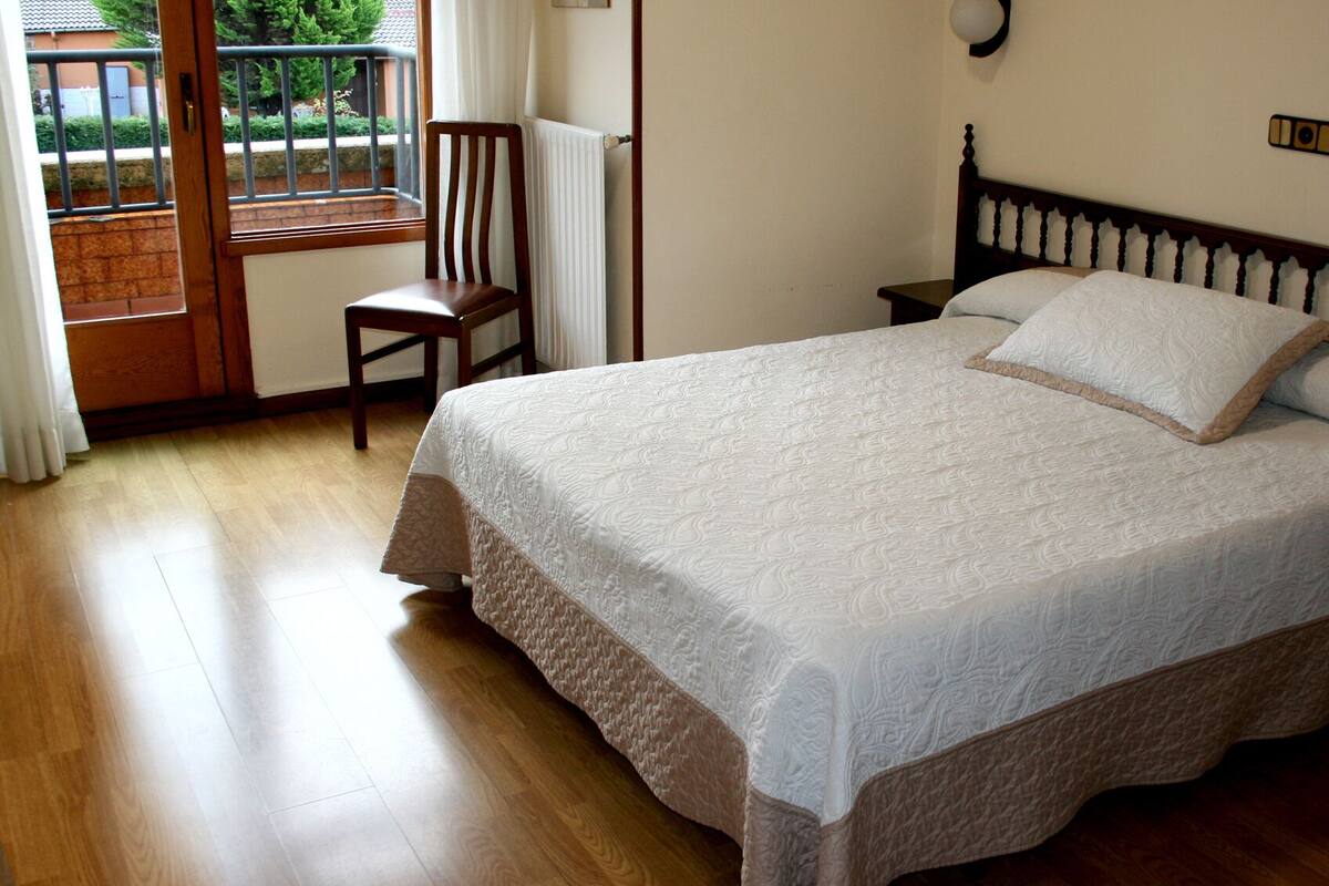 A bedroom with a large bed, wooden floor, a chair, a radiator, and a view of the outside.