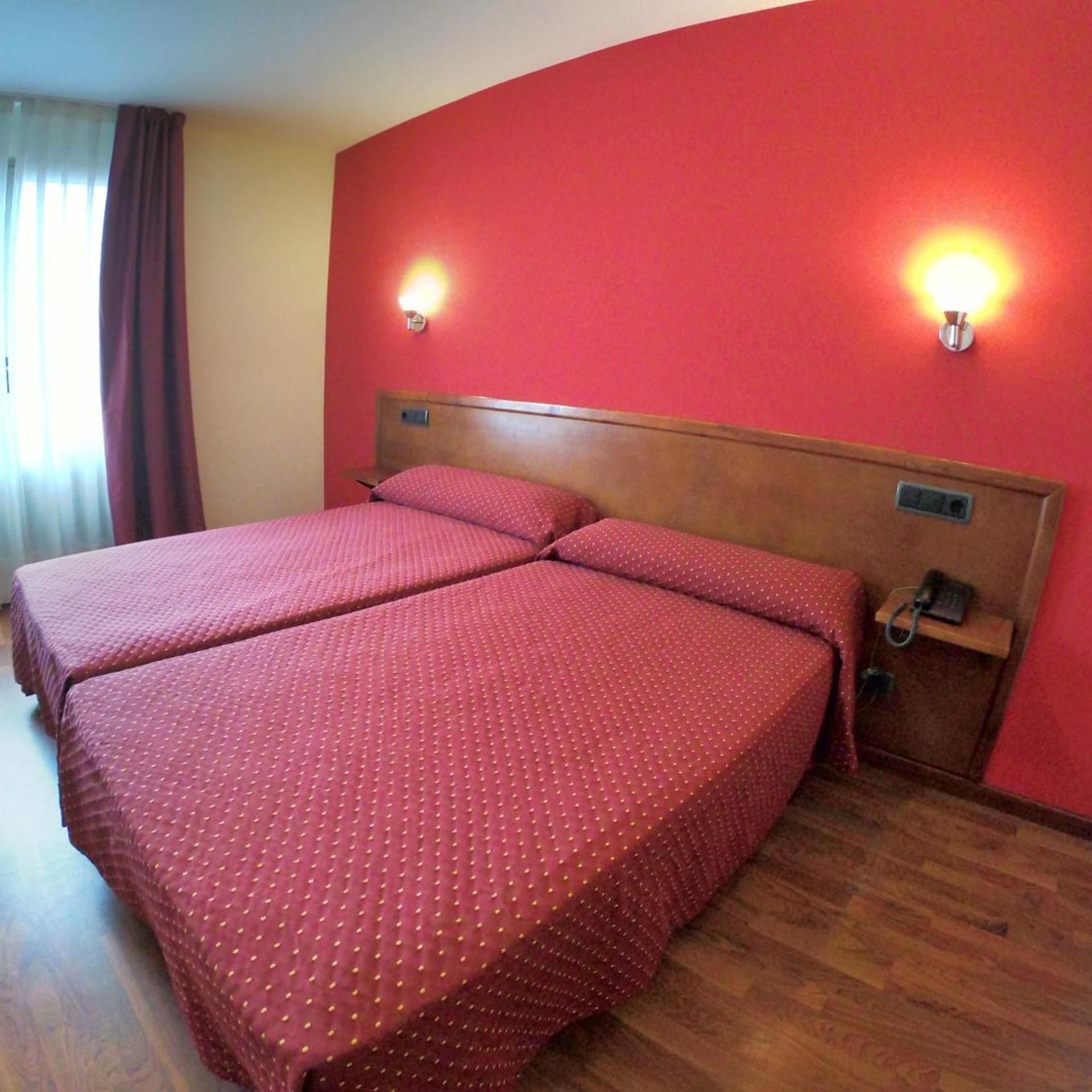Habitación de hotel con dos camas, cabecero de madera, paredes rojas y dos luces fijadas a la pared.