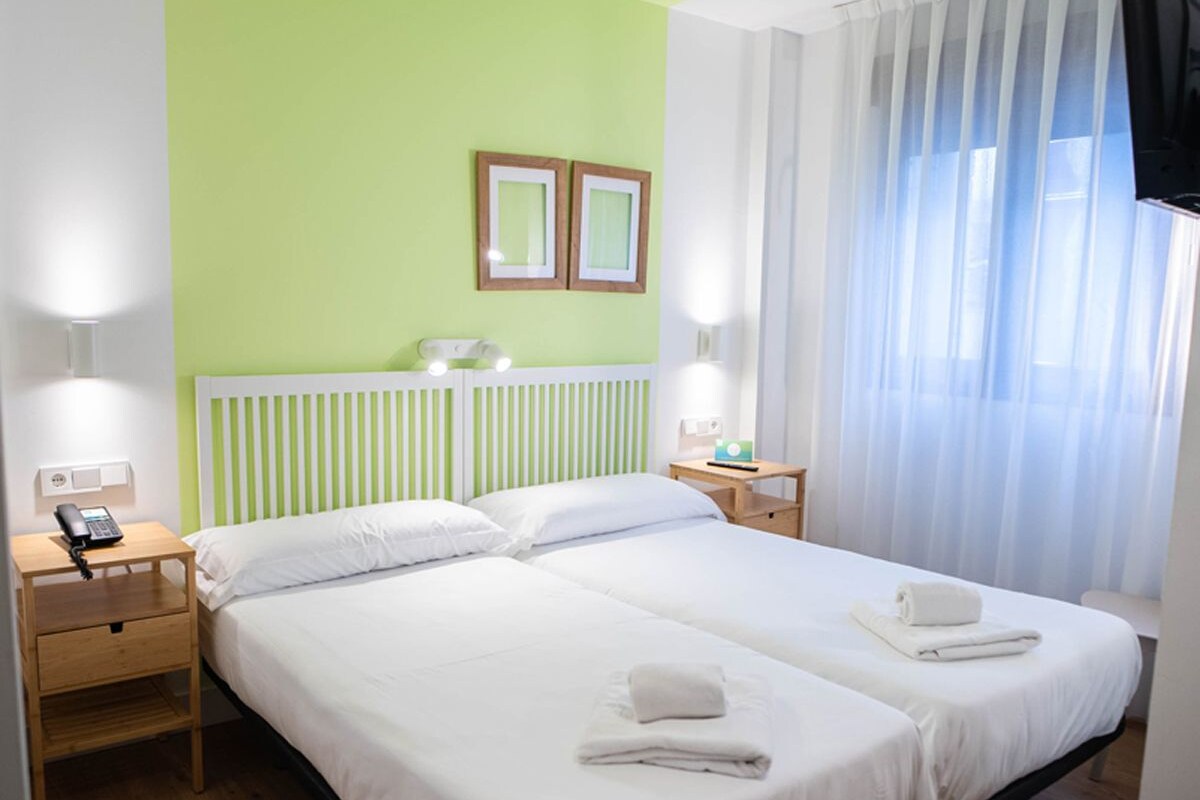 A hotel room with two beds, a bedside table with a lamp, a phone, and a window with sheer curtains.