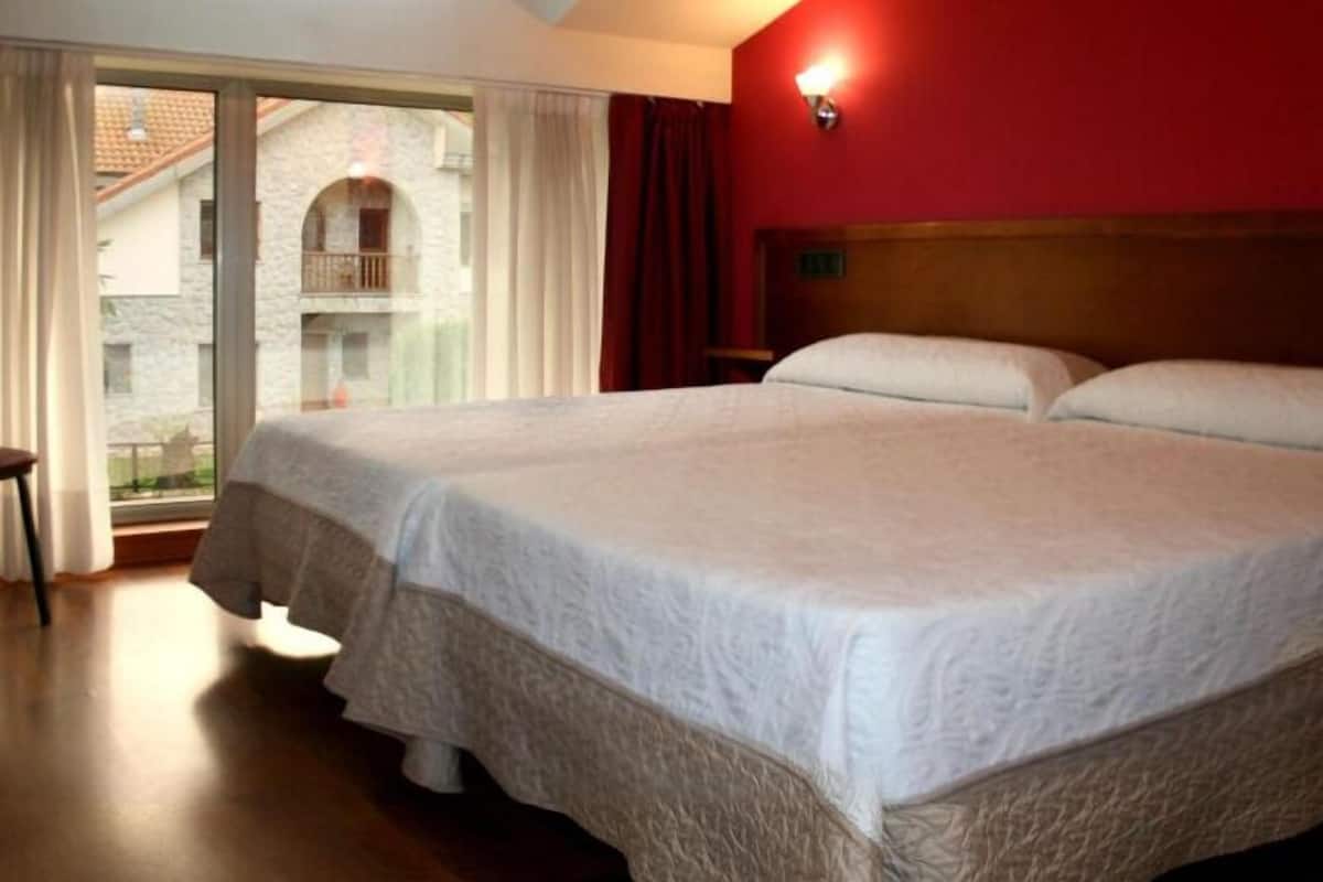 A double bed in a room with a wooden floor, a red wall, and a window with curtains.