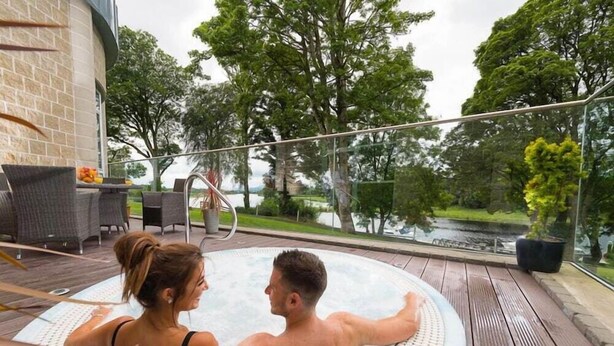 Ein Paar entspannt in einer Whirlwanne auf einer Holzterrasse mit Gartenmöbeln, mit Blick auf ein Gewässer und Bäume.