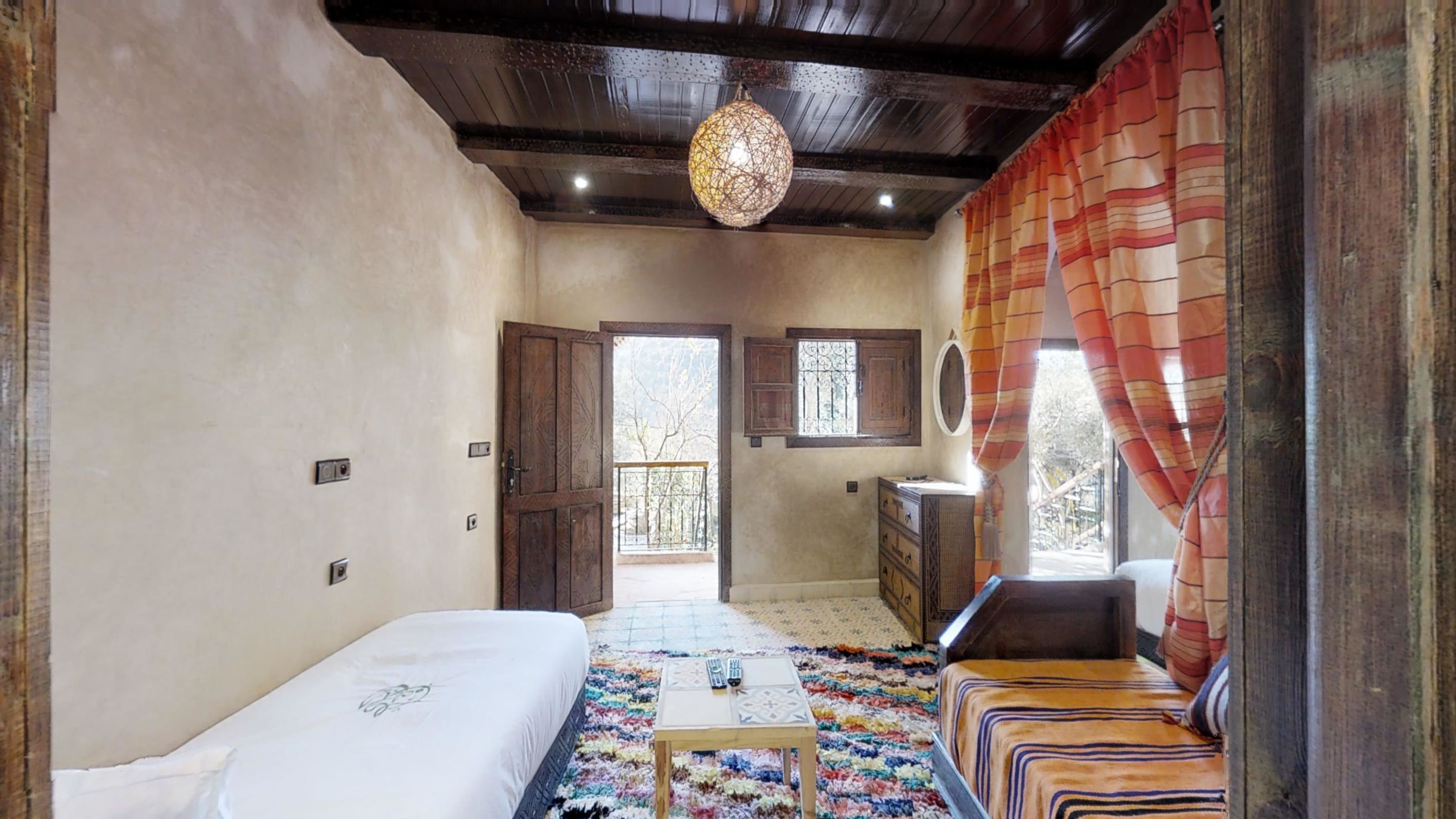 A room with a bed, a small table, a sofa, a hanging light fixture, and a view of the outside.