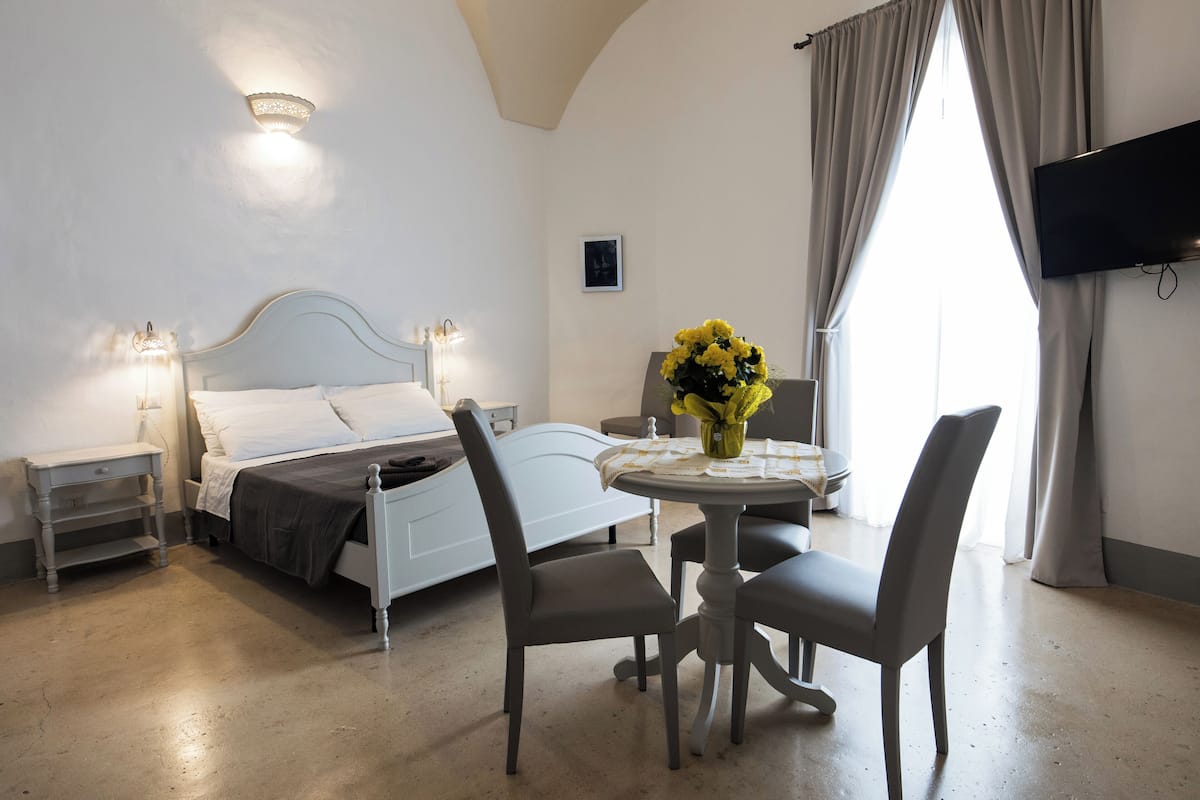 A bedroom with a bed, bedside tables, a TV, a dining table with chairs, and a window with curtains.