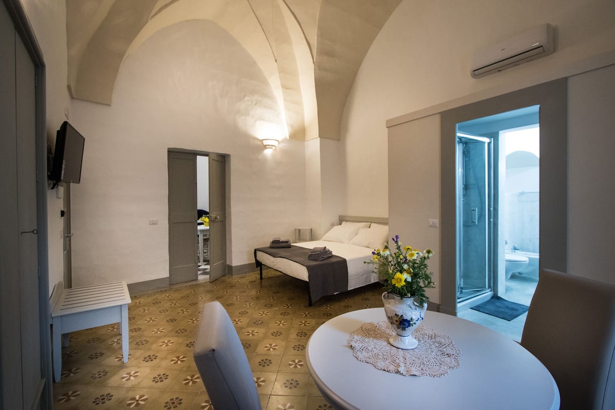 A hotel room with a bed, a small table with a vase of flowers, a chair, and a view of the outside.