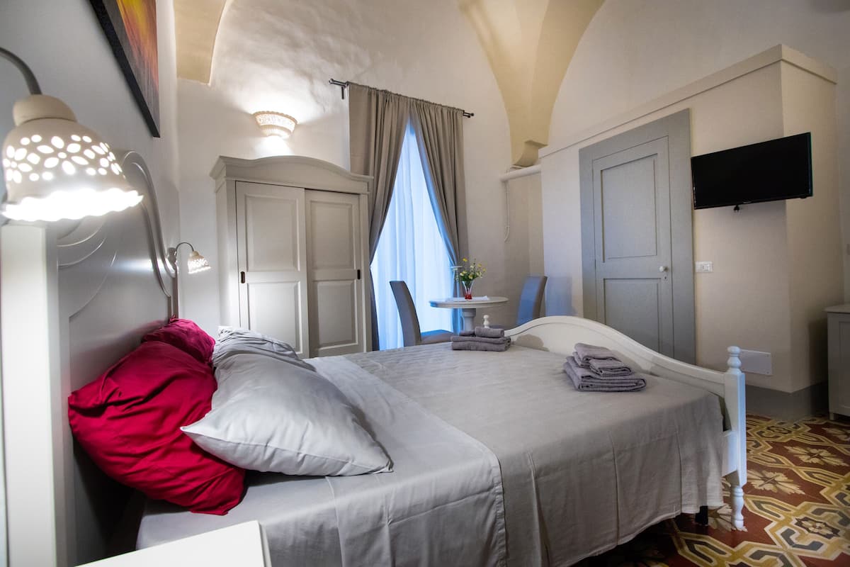 A bedroom with a bed, a nightstand, a lamp, a TV, and a window with curtains.