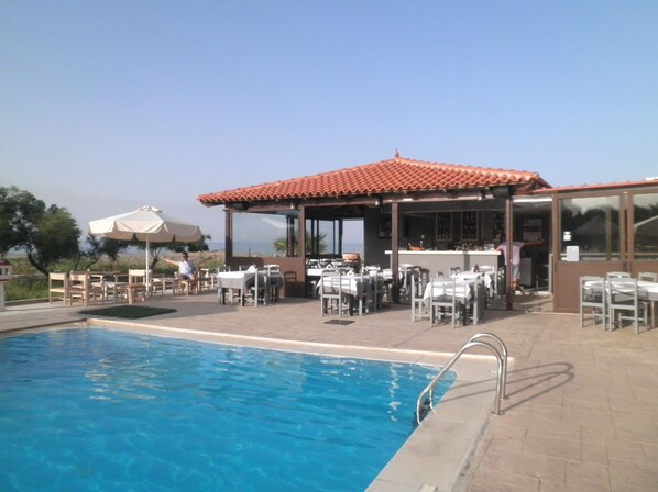 A poolside area with tables and chairs, a covered seating area, and a view of the sea. - Haridimos Apartments (Platanias)