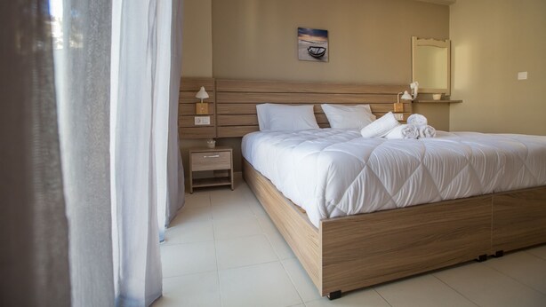 A neatly made bed with white linens, a wooden headboard, a bedside table with a lamp, and a painting on the wall. - Ilaeira rooms (Zakynthos)