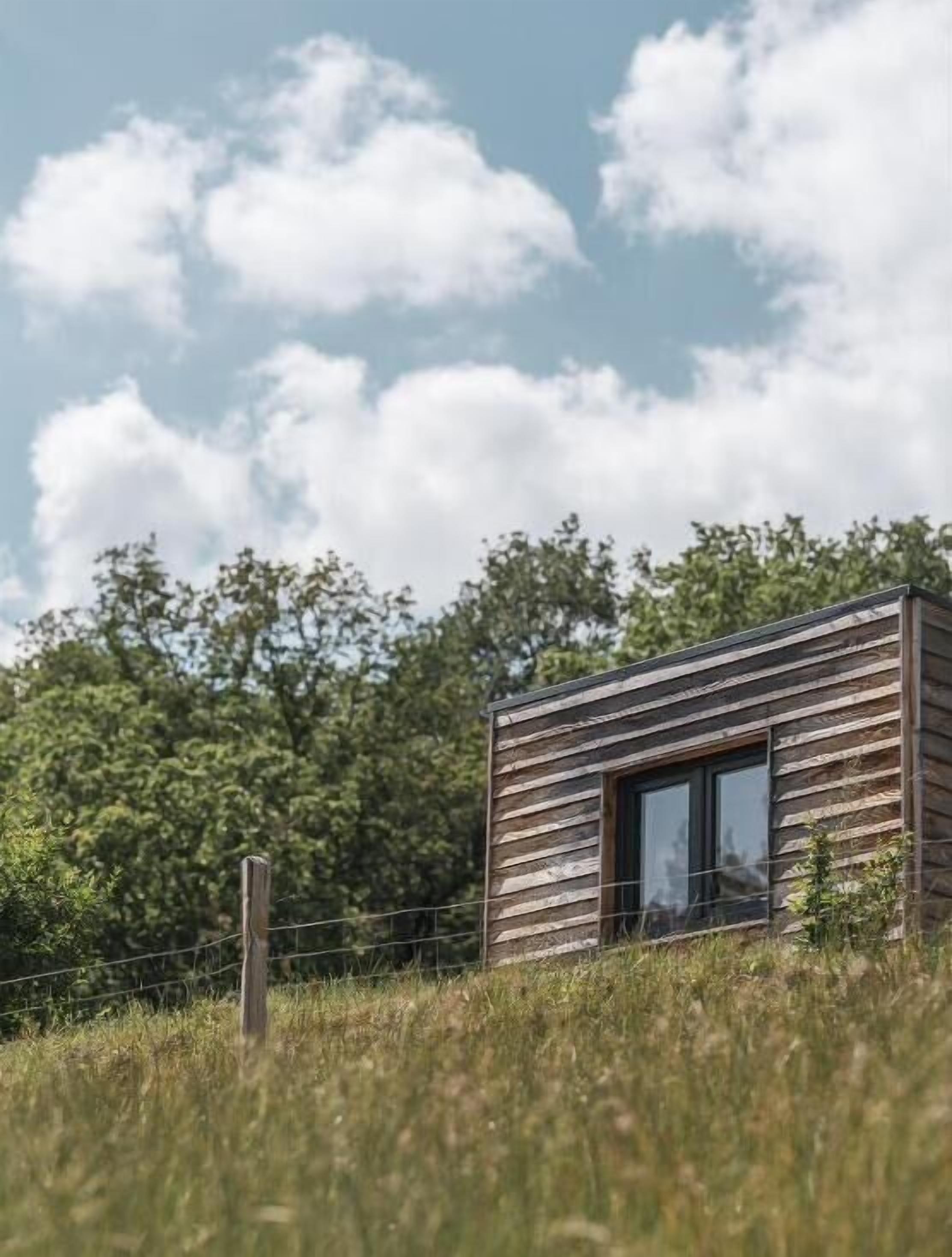 A small wooden cabin with a window, surrounded by a fence and grass, set against a backdrop of trees and a partly cloudy sky.